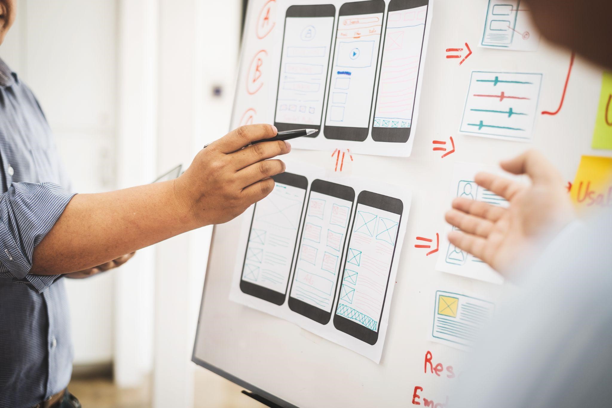 Designer pointing at hand-drawn mobile app wireframes pinned to a whiteboard during a planning discussion