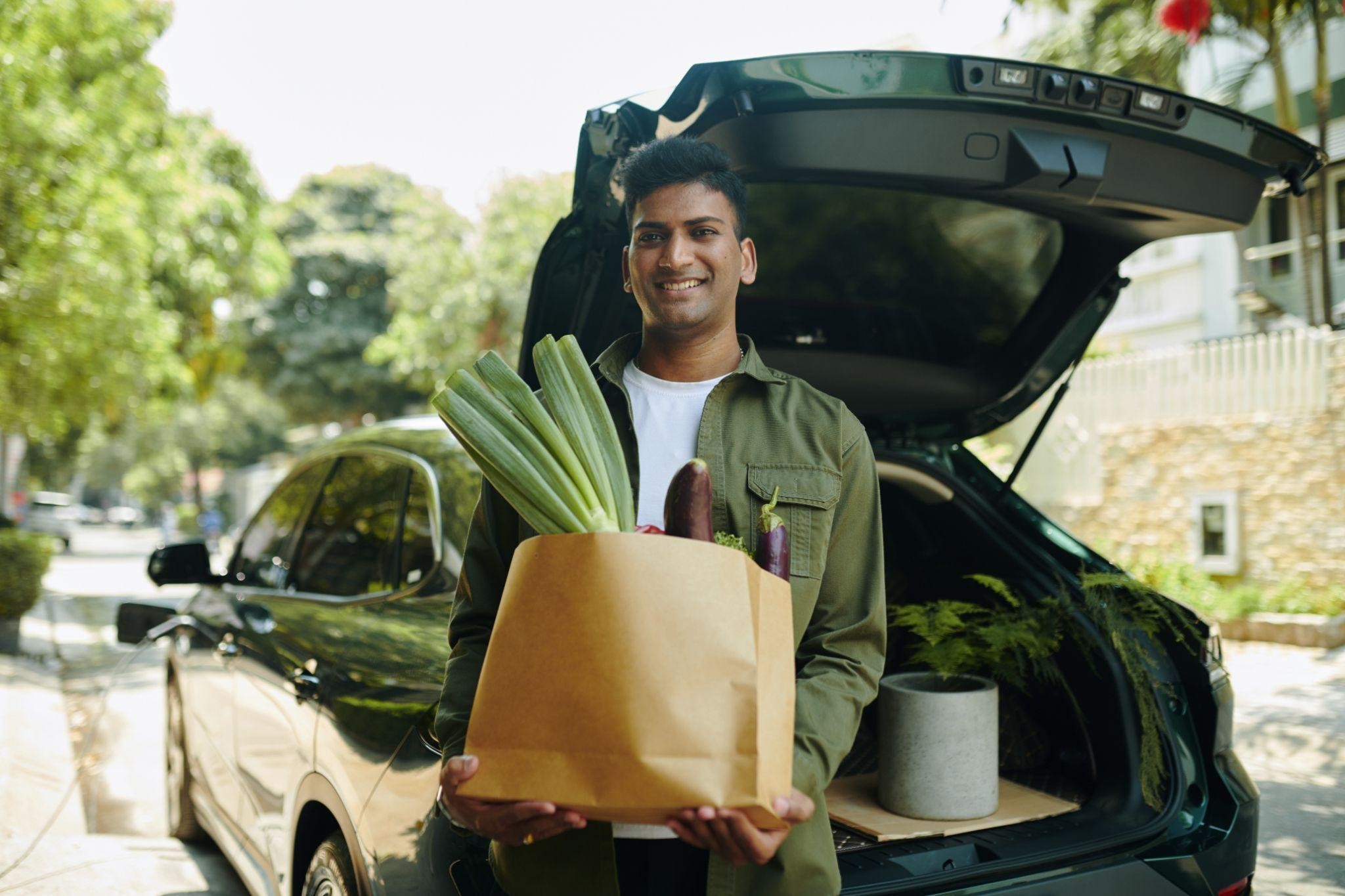 Smiling man holding a bag of fresh vegetables next to an open car trunk outdoors