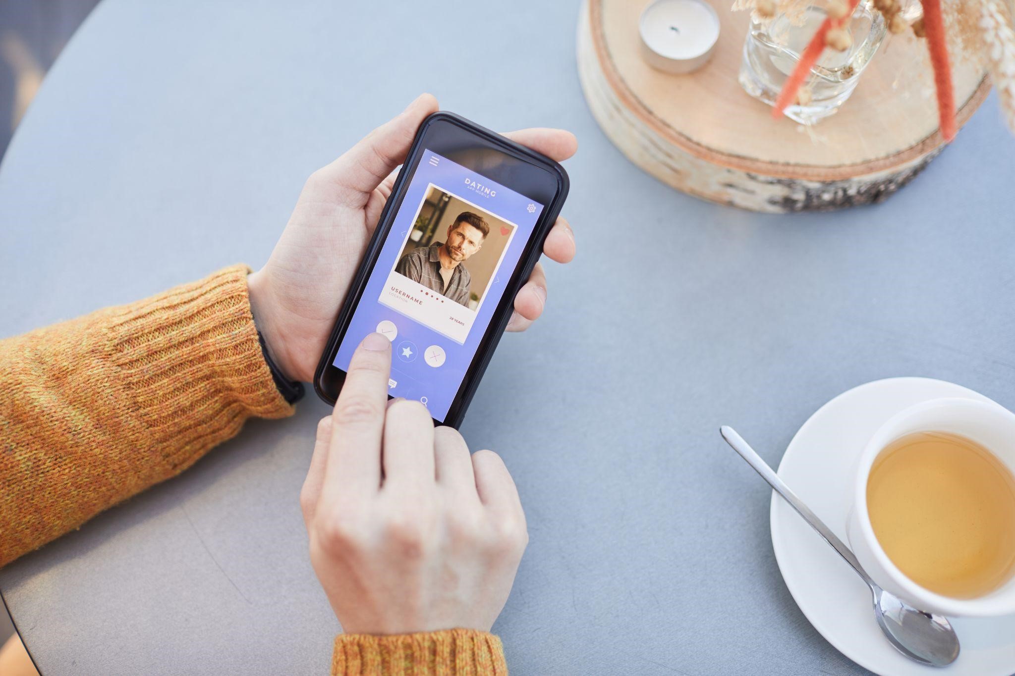 Overhead view of hands browsing a dating app profile on a smartphone next to a cup of coffee at a cafe table
