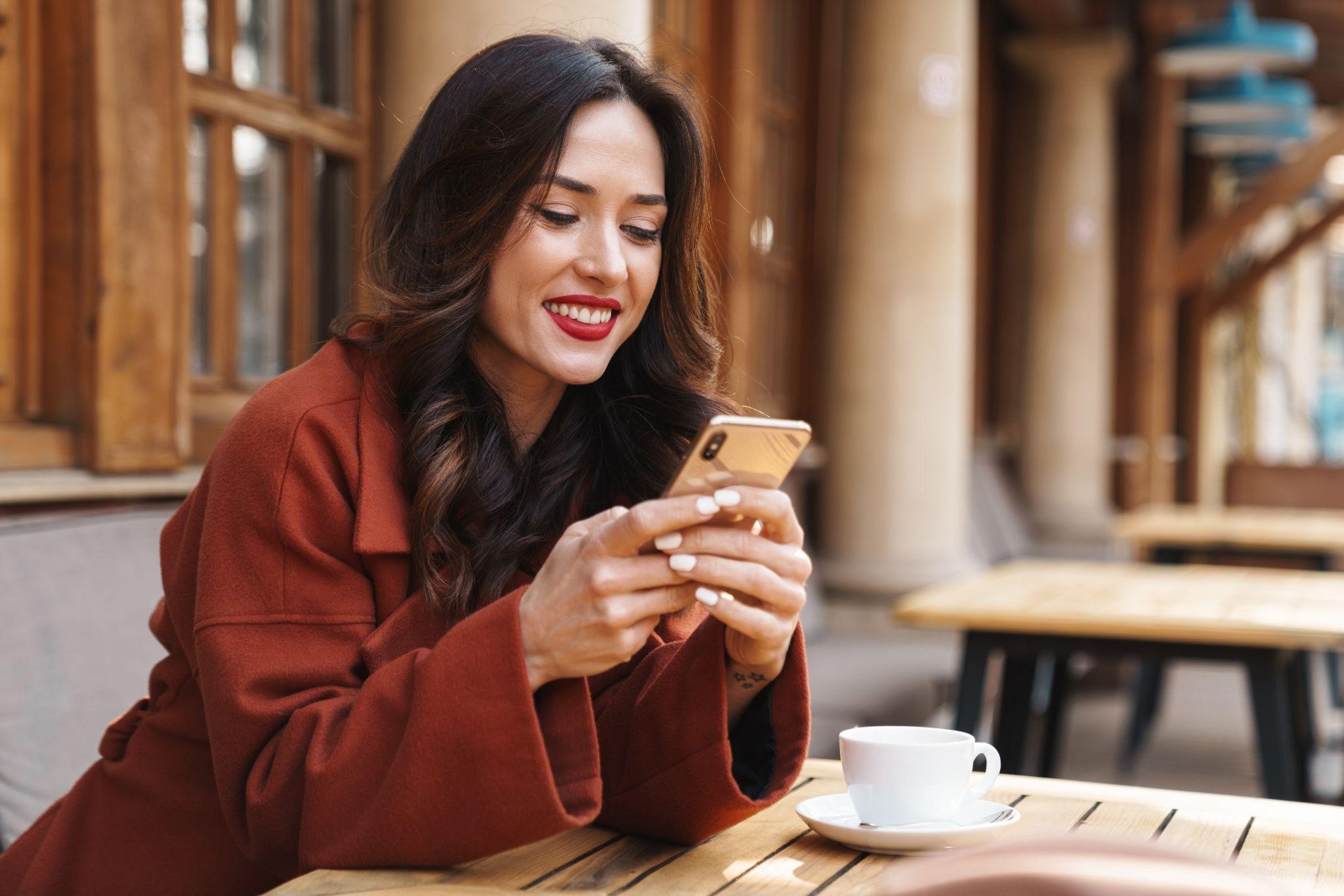 Smiling woman using her smartphone at an outdoor cafe table with a cup of coffee nearby
