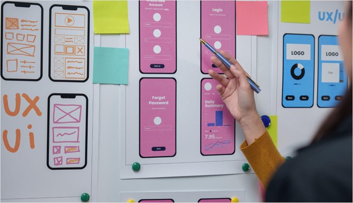 Designer pointing to a whiteboard covered in mobile app wireframes and UI mockups, with sticky notes and UX sketches pinned around them