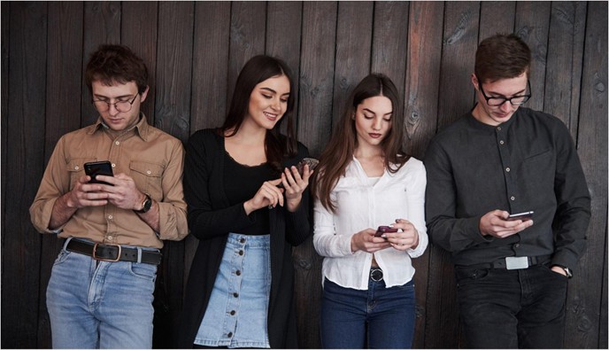 Four young adults standing against a wood wall each looking down at their smartphones